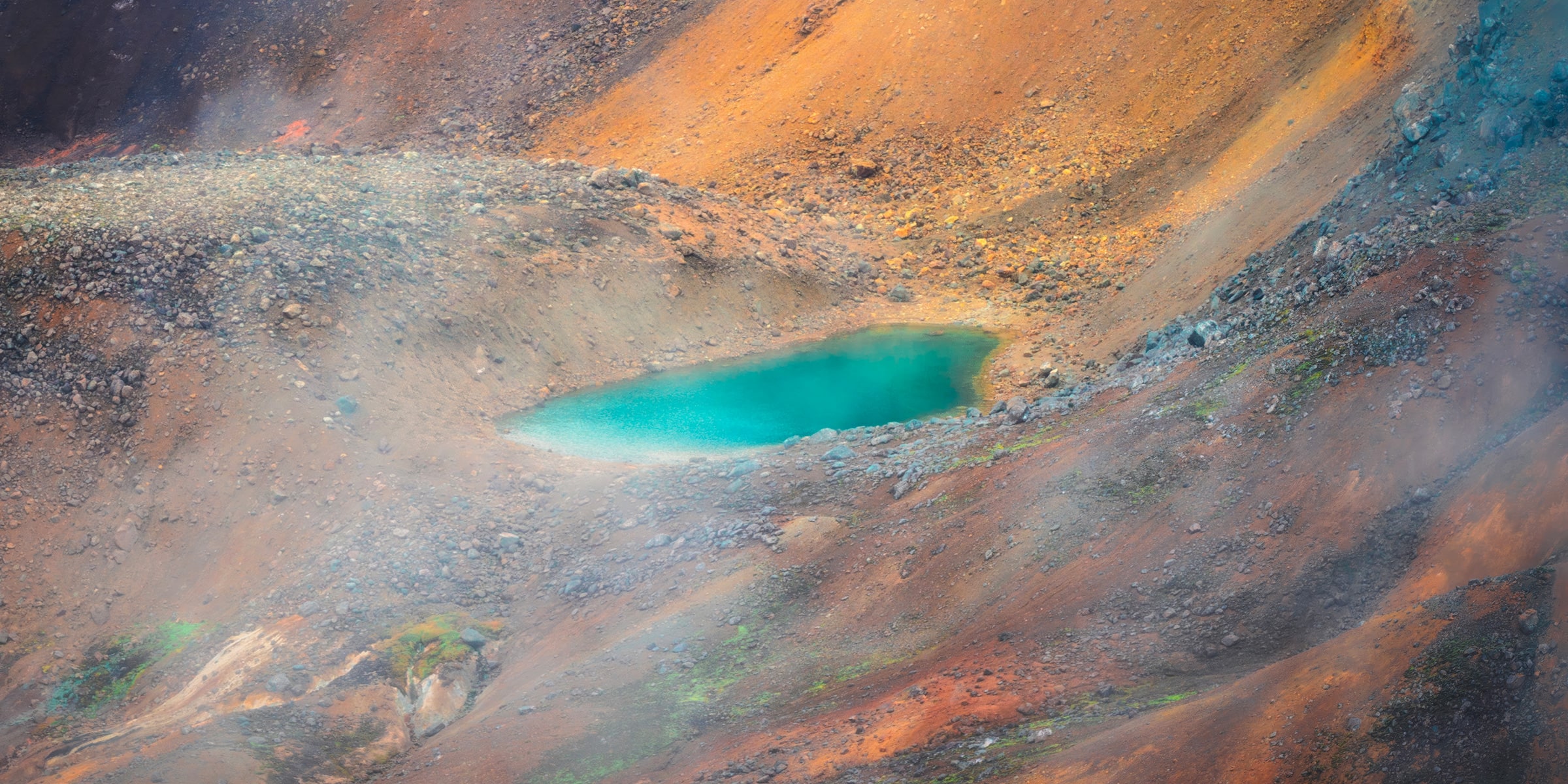 Aerial panoramic photograph of a turquoise geothermal pool surrounded by copper, amber, rust, and charcoal volcanic mountains in the Icelandic Highlands, with wisps of steam rising across the mineral-rich terrain.