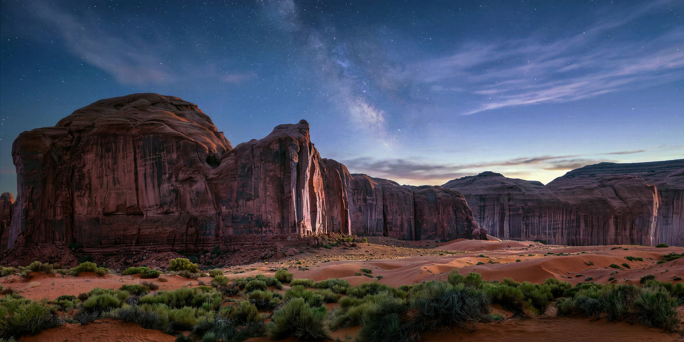 Fine art landscape photograph of Monument Valley by Sarah L. Glabman: towering red sandstone cliffs and softly lit desert dunes beneath a blue twilight sky, with a subtle band of the Milky Way rising above the rock formations.