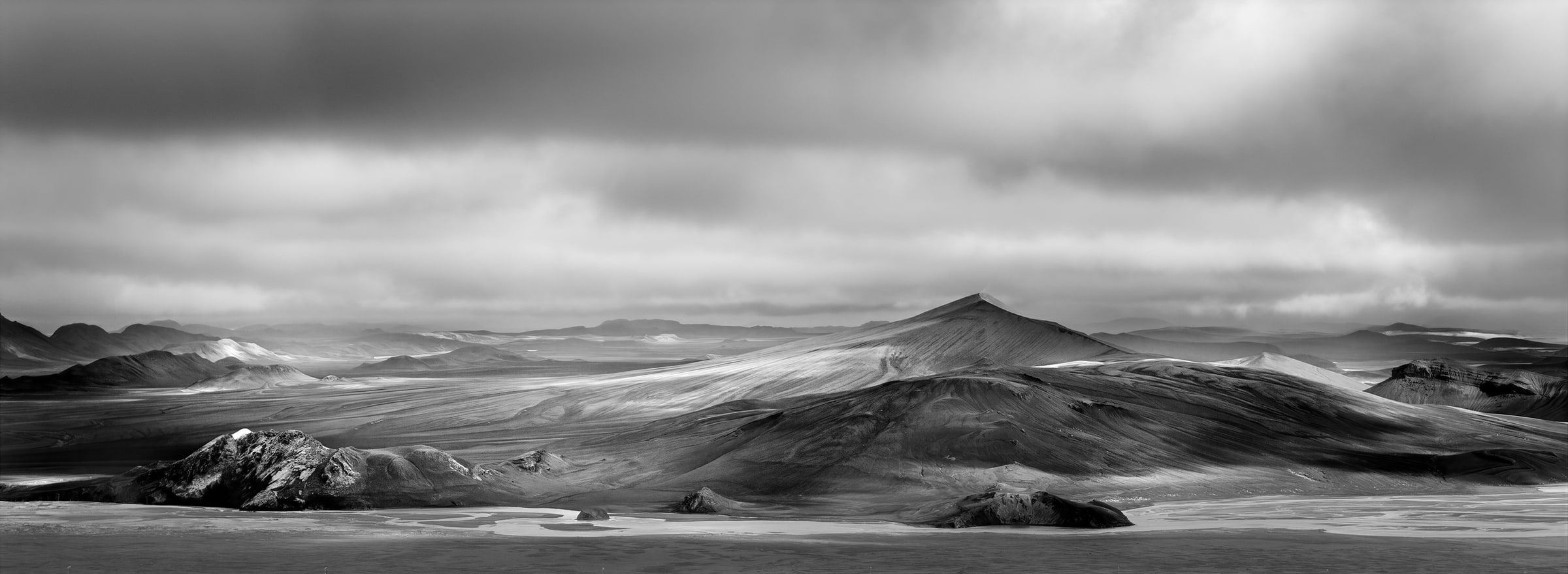 Black and white panoramic aerial photograph of Iceland’s remote volcanic mountains beneath dramatic storm clouds, created from dozens of stitched drone images.