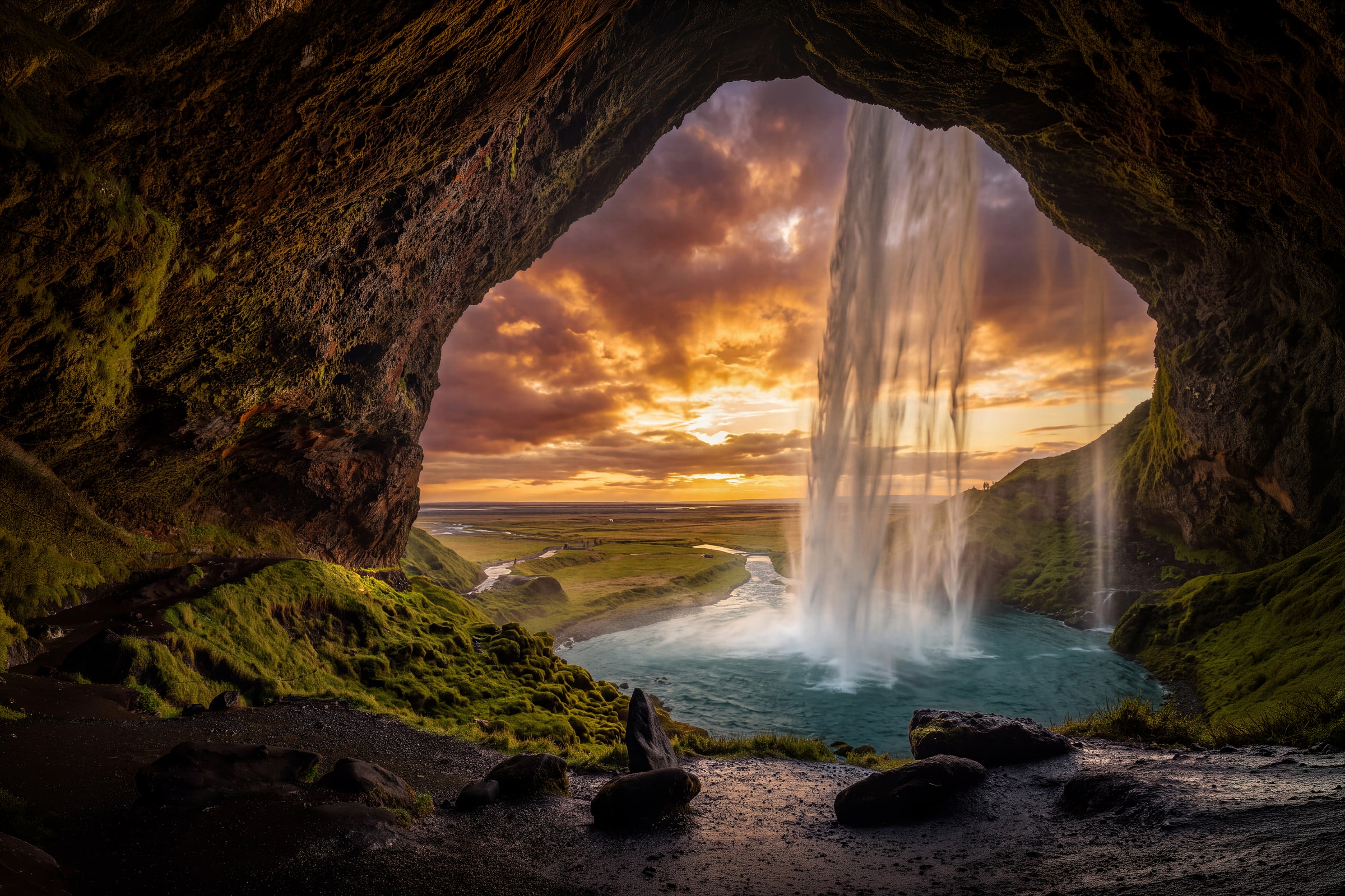 Fine art photograph of Seljalandsfoss waterfall in South Iceland by Sarah L. Glabman, taken from inside the volcanic cavern behind the falls, with a curtain of water dropping into a turquoise pool, green moss, and a winding river beneath a golden amber sunset.