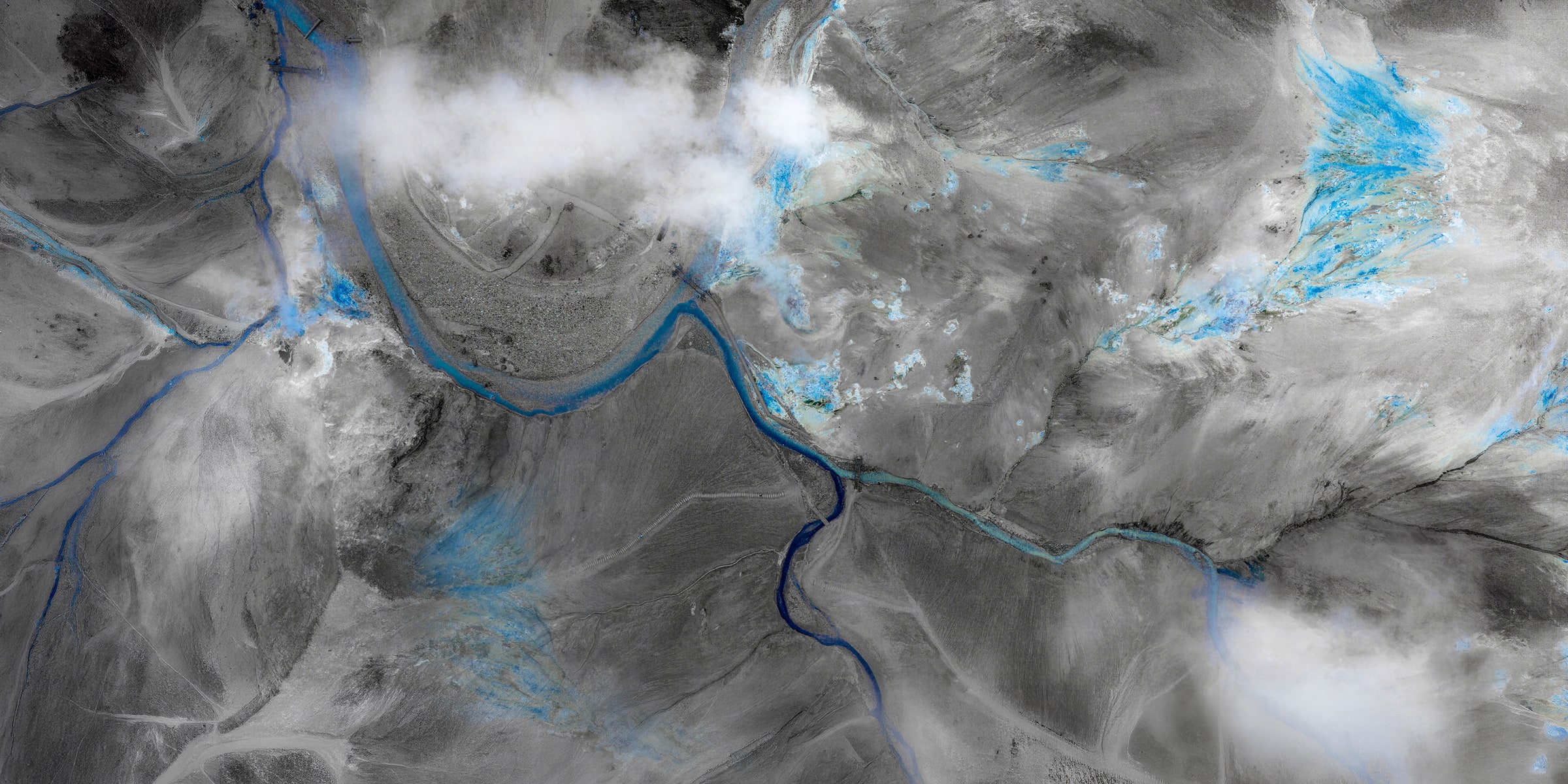 Aerial fine art photograph of Iceland’s geothermal highlands with winding sapphire-blue waterways across textured black-and-white volcanic terrain and drifting clouds.