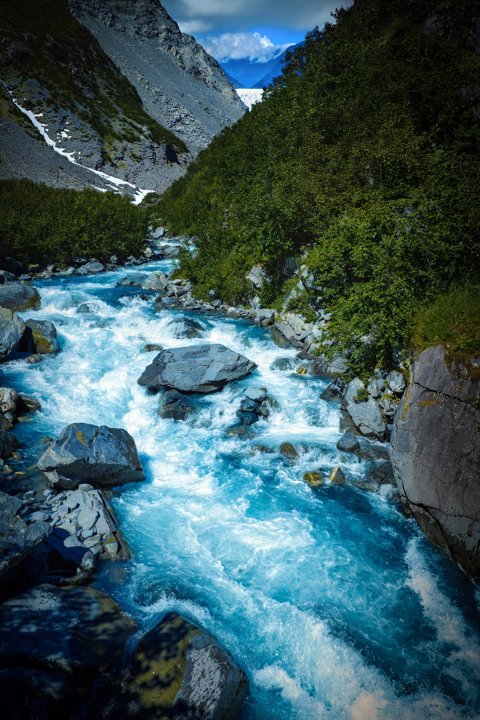 Aerial fine art photograph of a vivid turquoise glacial river rushing through a rocky mountain valley near Whittier and Prince William Sound, Alaska, surrounded by dense green vegetation with distant glacier ice visible between the mountains in the Chugach region.