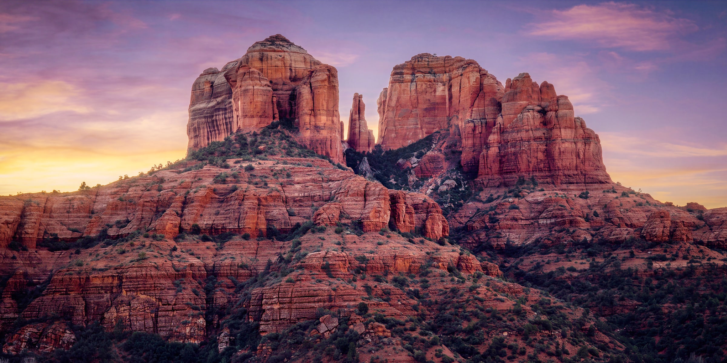 Panoramic sunrise photograph of Cathedral Rock in Sedona, Arizona, by Sarah L. Glabman: towering red sandstone formations above rugged desert terrain beneath a sky of lavender, pink, peach and golden light.