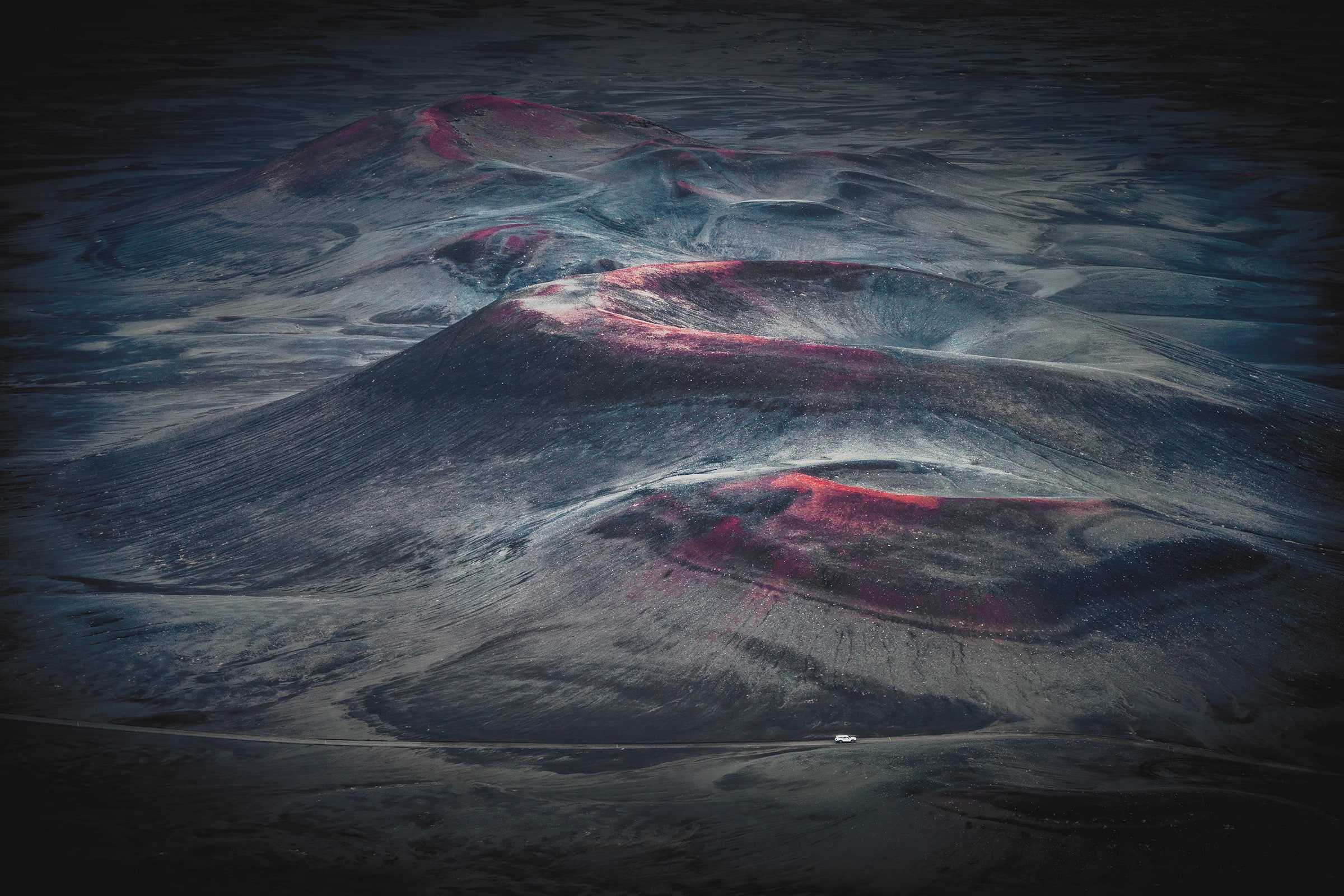 Drone photograph of sculptural charcoal volcanic hills in Iceland streaked with crimson and magenta tones, with a tiny white vehicle on a road below revealing the immense scale.