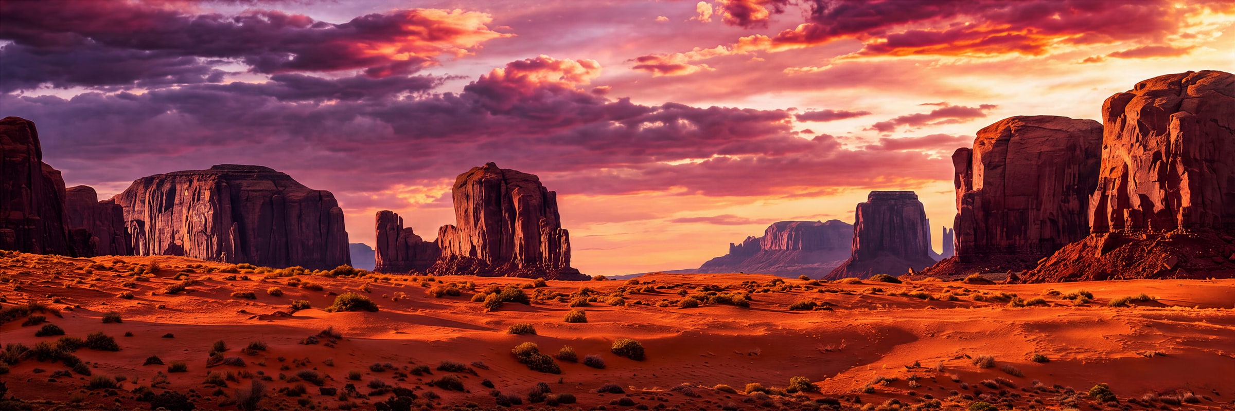 Panoramic fine art photograph of Monument Valley by Sarah L. Glabman: towering red sandstone buttes and mesas above a glowing orange desert floor beneath a sunset sky of deep purple, magenta, pink and golden clouds.