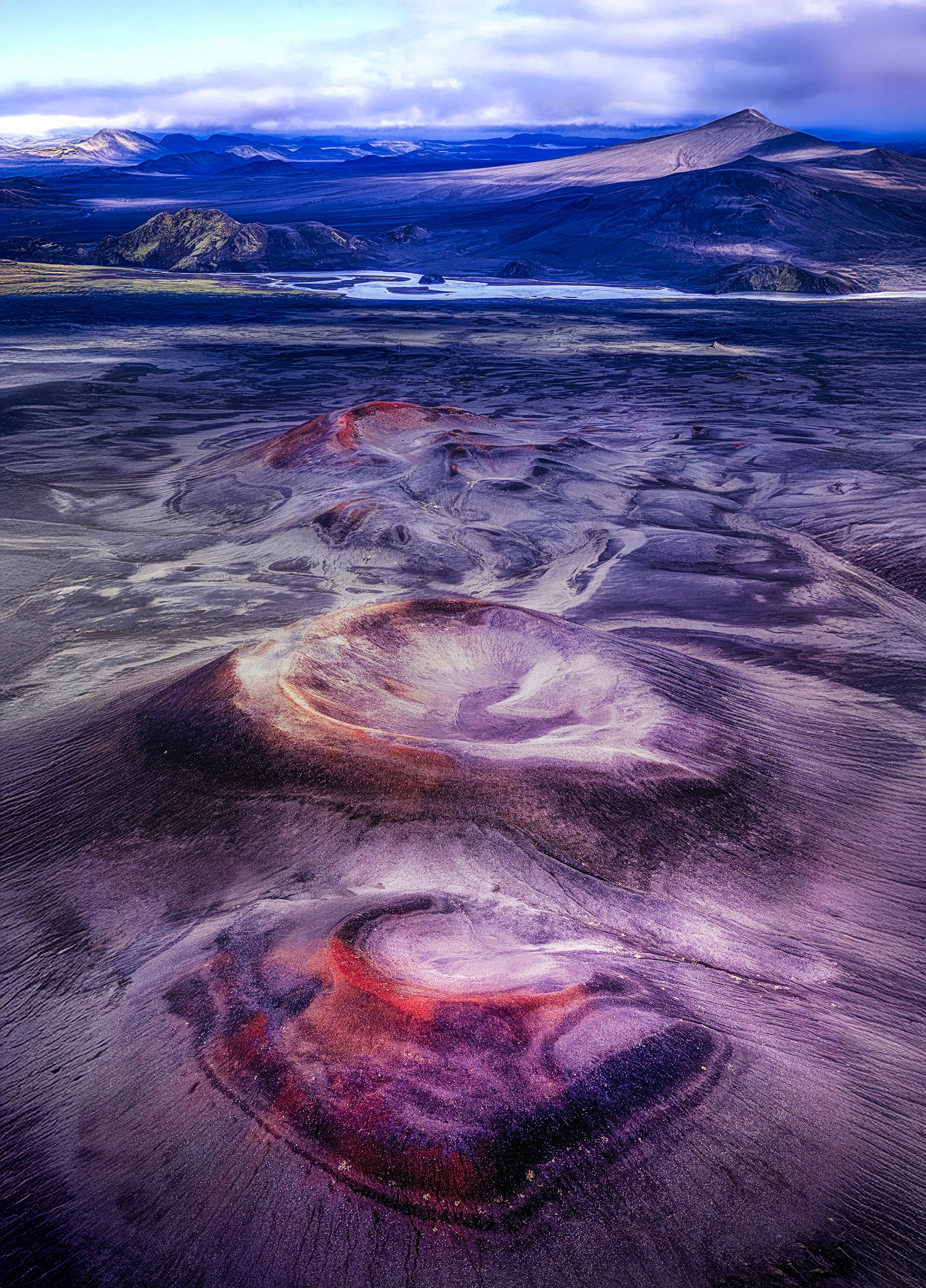 Fireborn by Sarah L. Glabman, a vertical aerial fine art photograph of Iceland's volcanic landscape featuring three colorful volcanic formations in shades of crimson, violet, rust and black, surrounded by vast lava terrain, braided waterways and distant Icelandic mountains beneath a dramatic sky.