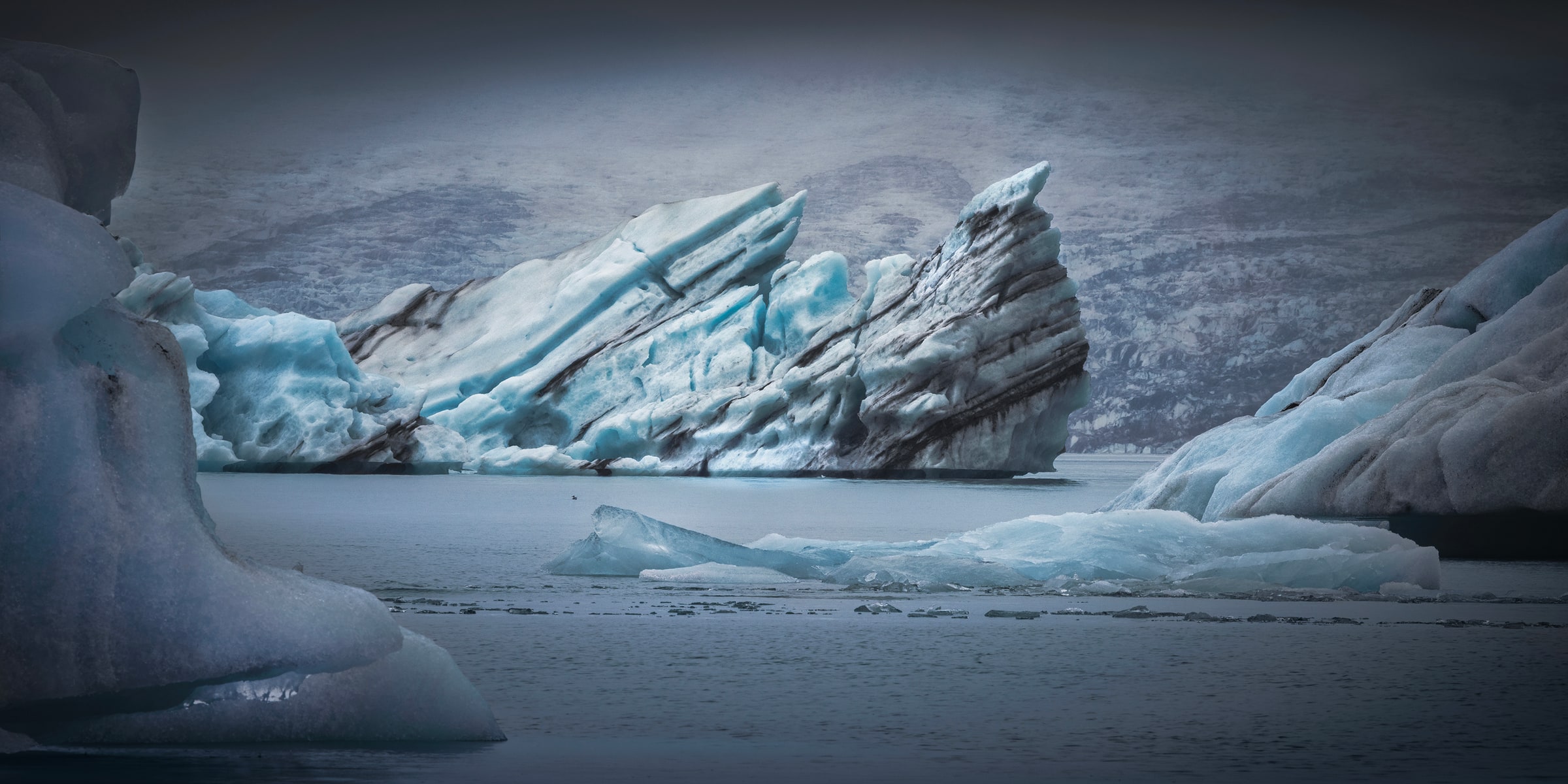 Massive sculpted ice formations frame towering turquoise-blue icebergs streaked with dark volcanic sediment at Jökulsárlón Glacier Lagoon, Iceland, beneath a moody gray sky.