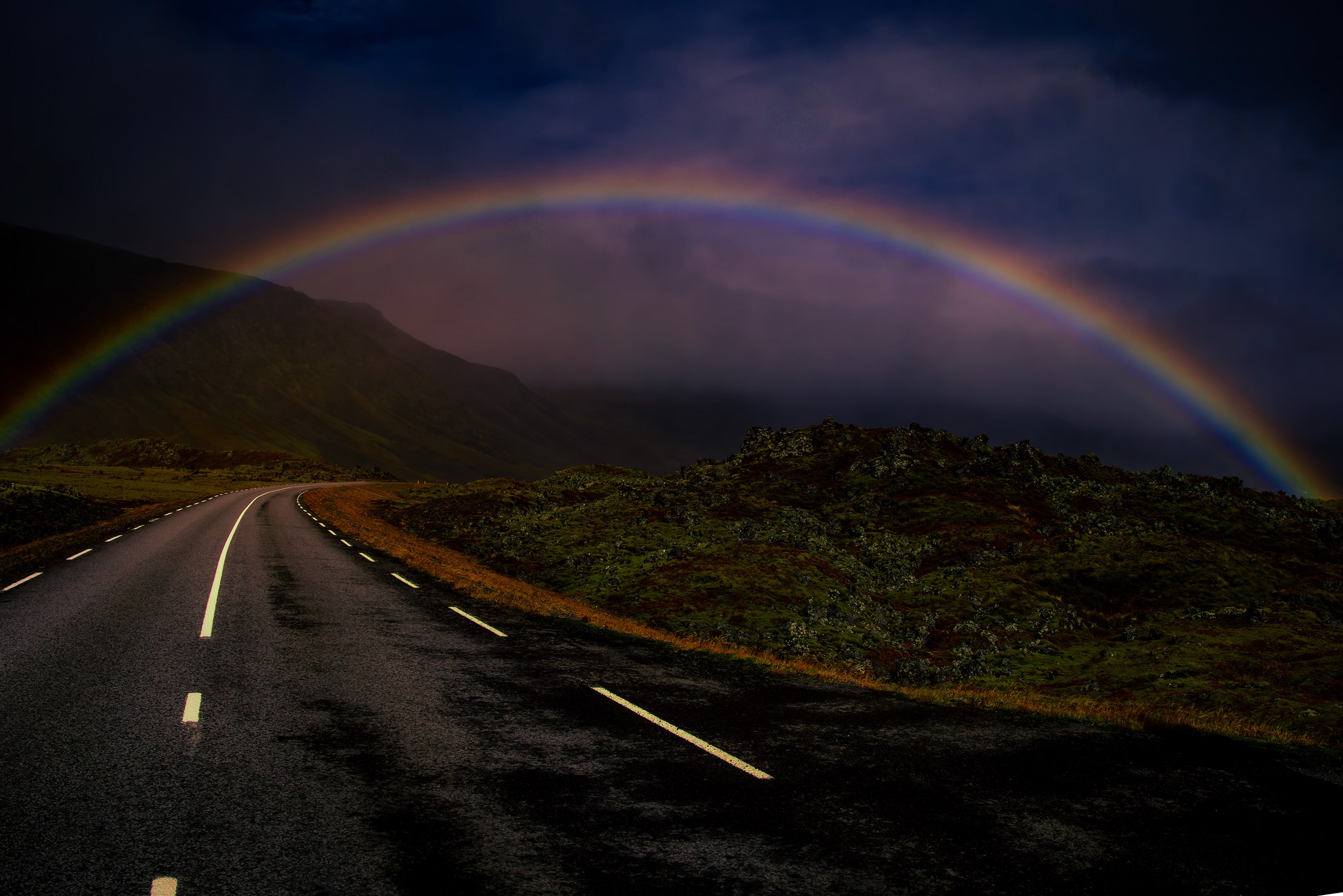 A full rainbow arching over an Icelandic valley and the road beneath dark storm clouds