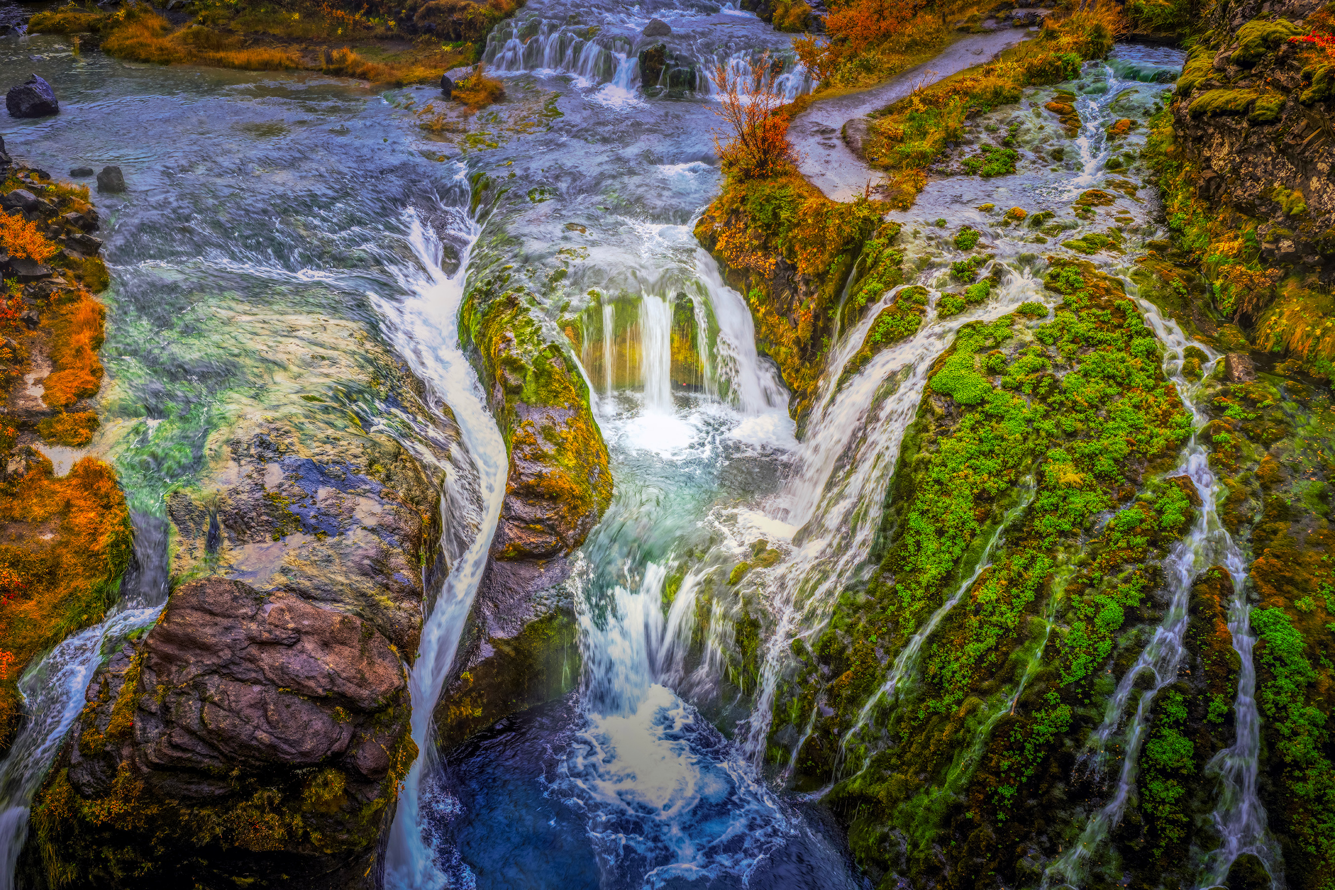 The Secret Path: aerial view of Gjáin in the Þjórsárdalur Valley, South Iceland, blue water spilling through moss-covered lava rock and autumn color