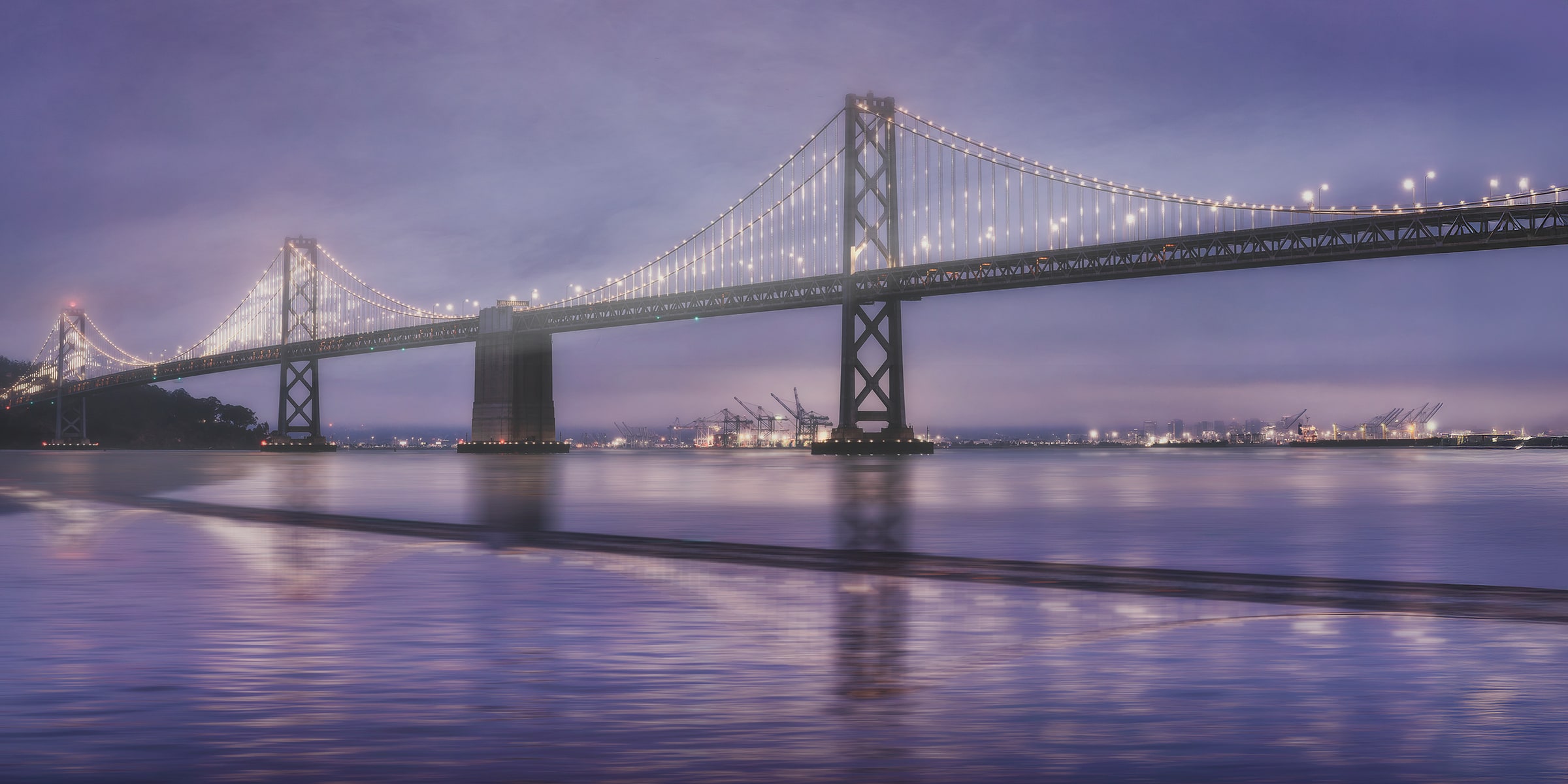 Fine art photograph of the illuminated San Francisco Bay Bridge during blue hour before sunrise, surrounded by misty San Francisco fog beneath lavender and violet skies, with softly reflected light across the calm waters of the bay.