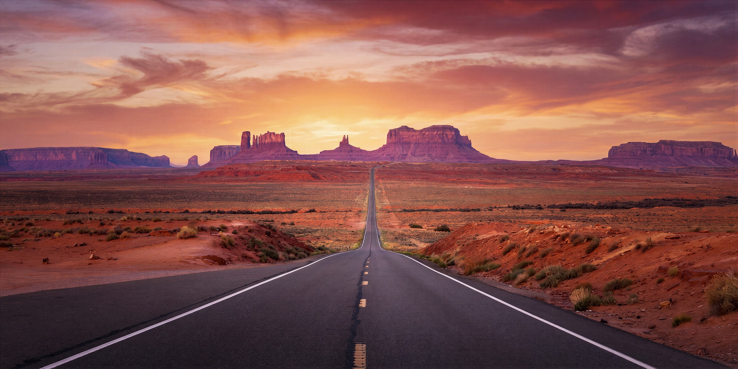 Panoramic photograph of the Forrest Gump Point highway near Monument Valley, Utah: a straight desert road leading toward red sandstone buttes beneath a sunset sky of crimson, orange, pink and violet clouds.