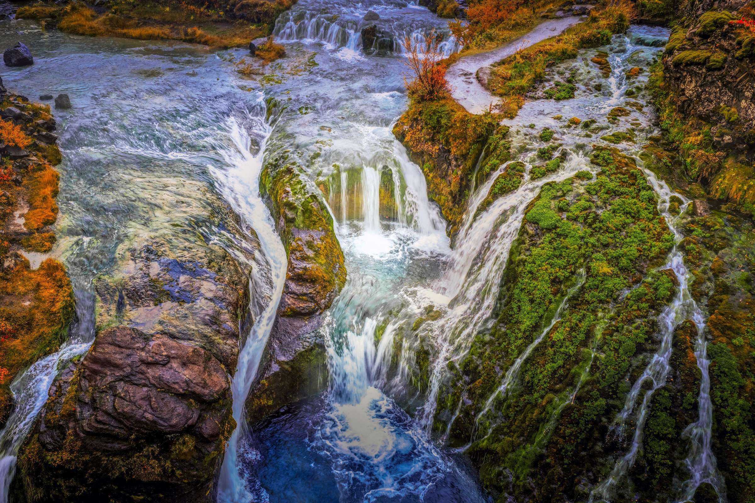 Aerial fine art photograph of Gjáin in the Þjórsárdalur Valley of South Iceland captured by Sarah L. Glabman, showing brilliant blue water cascading through moss-covered volcanic rock surrounded by vivid orange and gold autumn foliage. A pale winding pathway curves between the waterfalls, creating the appearance of a secret passage through the Icelandic landscape.