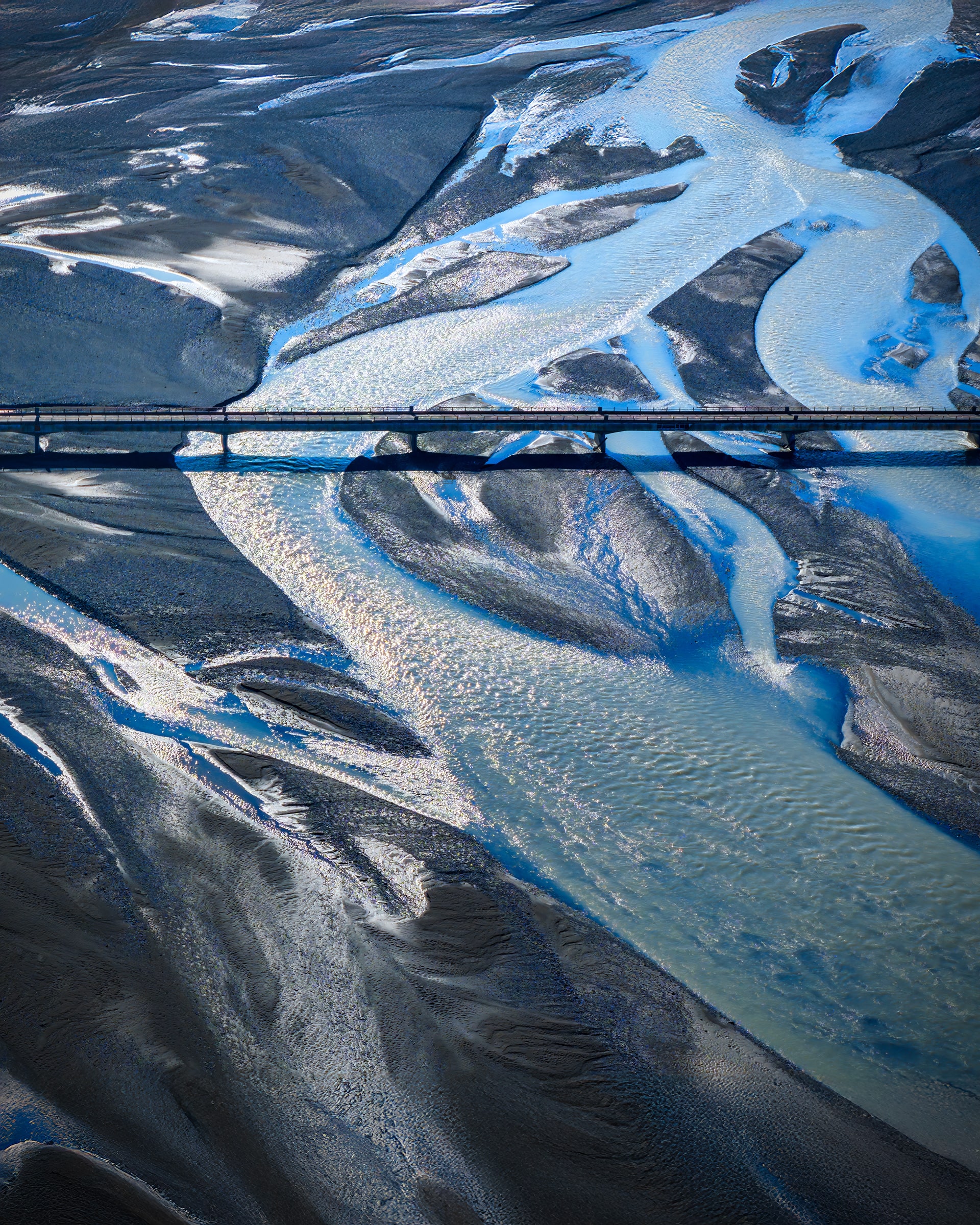 Threshold by Sarah L. Glabman is a fine art aerial photograph created by drone above Iceland, revealing a narrow bridge and roadway surrounded by expansive black volcanic lava fields. The dramatic Icelandic landscape creates a minimalist composition symbolizing transition, possibility, and the journey into a new chapter.