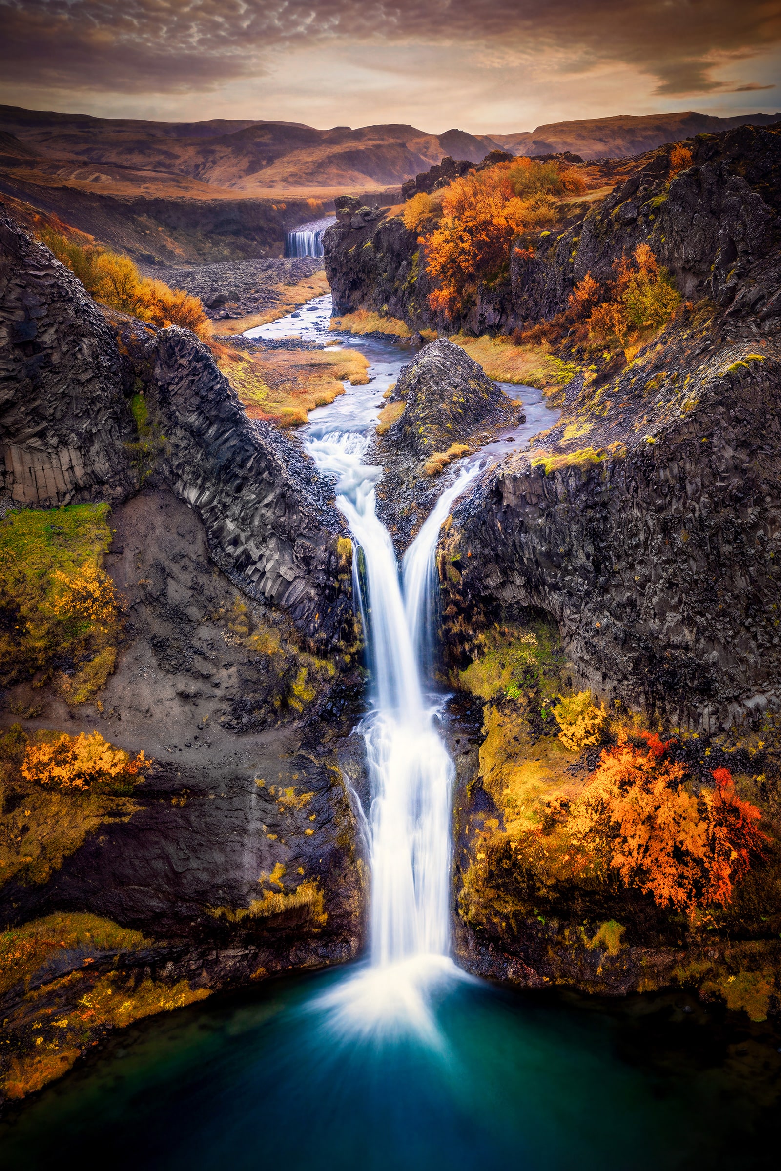 Aerial fine art photograph by Sarah L. Glabman of a waterfall in Gjáin Canyon, Iceland, falling between dark basalt cliffs and fiery orange autumn foliage into a turquoise pool, with a winding river and mountains beyond.