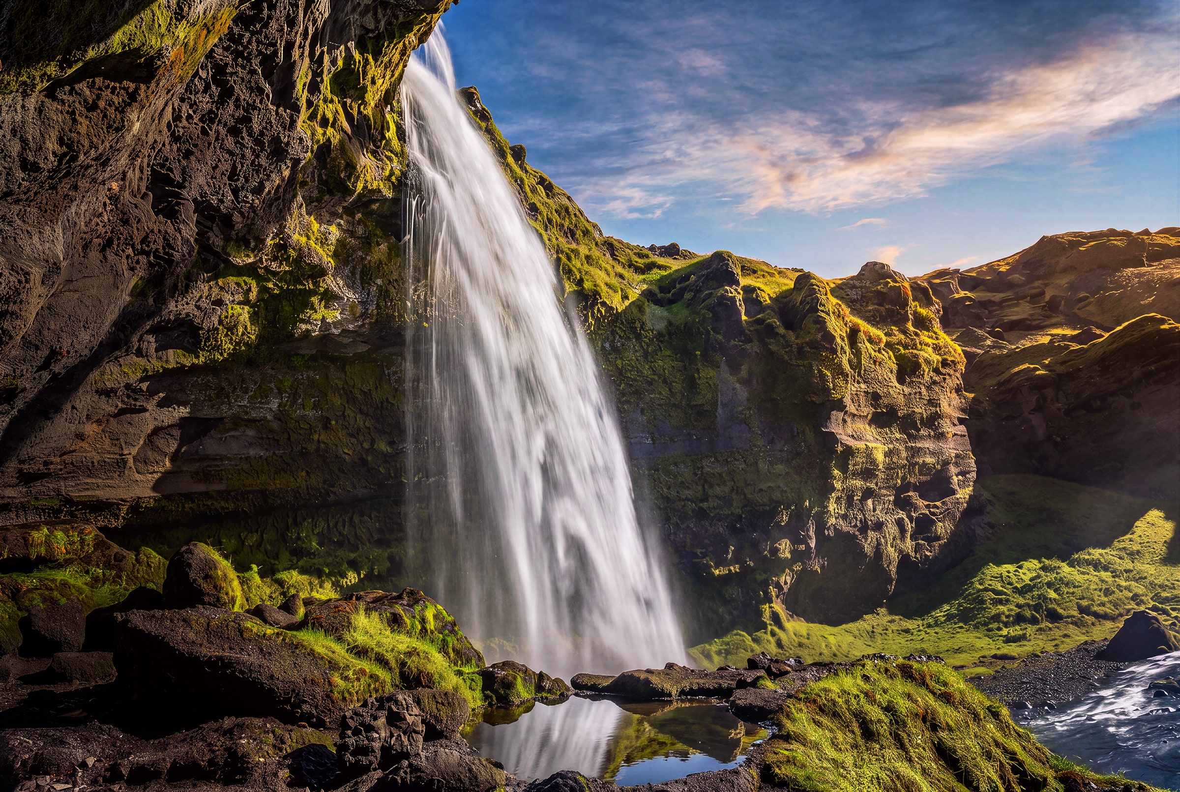Fine art photograph of Kvernufoss waterfall in South Iceland by Sarah L. Glabman, taken from beneath a dark volcanic rock overhang, with silky white water, vivid green moss, sunlit cliffs, blue sky and a small reflection pool in the foreground.